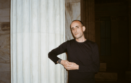 A portrait of graphic designer Filippos Fragkogiannis taken in Athens, October 2023. He is wearing a black long-sleeve shirt and leans against a classical marble column with a calm and composed expression. The background features an architectural setting with neoclassical elements, including stone walls and pillars, captured in warm, natural lighting that enhances the atmosphere. Filippos Fragkogiannis