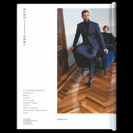 A stylish fashion editorial spread featuring a sharply dressed man in a navy-blue suit with a black turtleneck sweater, sitting elegantly on a modern dark blue velvet chair. He wears polished black leather shoes and exudes confidence as he gazes into the distance. In the background, another man, also in a dark suit, lounges on a matching blue velvet couch. The luxurious setting has a rich wooden herringbone-patterned floor, a pale blue wall with intricate molding, and a wooden door. The left page of the spread displays the years 