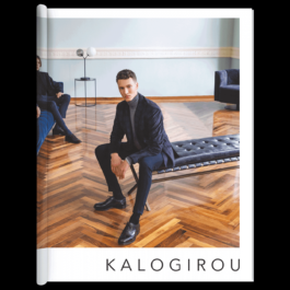 A fashion editorial cover featuring a well-dressed man sitting on a sleek black leather bench in a luxurious room with a herringbone-patterned wooden floor. He wears a dark blue suit with a turtleneck sweater and polished black leather shoes, gazing directly at the camera. In the background, another elegantly dressed man sits on a blue velvet couch, partially visible. The room has soft blue walls with white molding, and minimalistic furniture, including a modern floor lamp and a round side table. The magazine cover prominently displays the brand name 