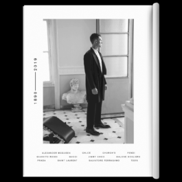 A black and white photograph of a man in a tailored black coat and formal attire standing in an elegant room with classic architectural details. He gazes out of a large window, with a marble bust statue placed on a pedestal behind him. The floor features a geometric tile pattern, and a modern black leather chair is partially visible in the foreground. The page design includes a vertical timeline reading 